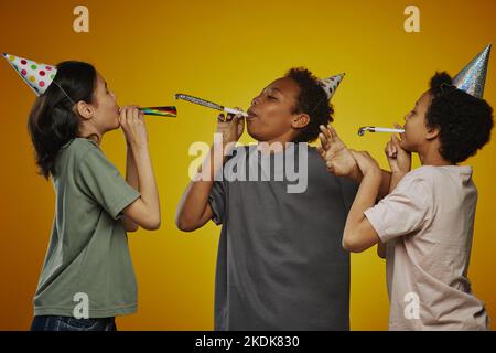 Gruppo di tre bambini pre-teen interculturali in t-shirt e cappellini di compleanno che soffiano fischietti mentre si diverte e si diverte Foto Stock