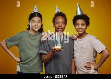 Tre giovani amici multiculturali in berretti di compleanno e casualwear in piedi di fronte alla macchina fotografica, mentre il ragazzo tiene la torta con le candele Foto Stock