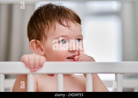 Il bambino del toddler si alza pensare nella greppia, ritratto di primo piano. Il bambino morde le unghie sulle dita, faccia del bambino. Kid è un ragazzo di un anno e quattro mesi Foto Stock