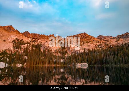 Alpine Lake, situato nella Sawtooth Wilderness dell'Idaho, visto in una mattinata d'estate all'alba, con luce del sole arancione che illumina Packrat e Monte Verita Peak Foto Stock