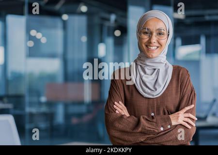 Ritratto di una donna d'affari felice e di successo in hijab, lavoratore d'ufficio sorridente e guardando la macchina fotografica a braccia incrociate, lavorando all'interno di un ufficio moderno. Foto Stock