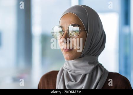 Primo piano ritratto di una donna commerciale musulmana che indossa hijab e occhiali, lavoratore in ufficio che guarda fuori finestra pensando alla decisione, capo femmina all'interno dell'ufficio. Foto Stock