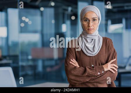 Donna d'affari seria e focalizzata in hijab e occhiali guardando la macchina fotografica a braccia incrociate, lavorando all'interno di un moderno edificio per uffici. Foto Stock