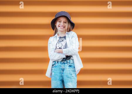 Piccolo ritratto ragazza vestita di moda sinceramente sorridente mentre guardando la fotocamera su sfondo arancione parete. Gente urbana che vive e una strada ogni giorno li Foto Stock