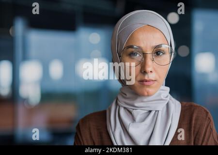 Primo piano ritratto di una donna di lavoro pensante di successo indossando hijab e occhiali, donna che guarda la macchina fotografica che lavora all'interno di un moderno edificio per uffici. Foto Stock