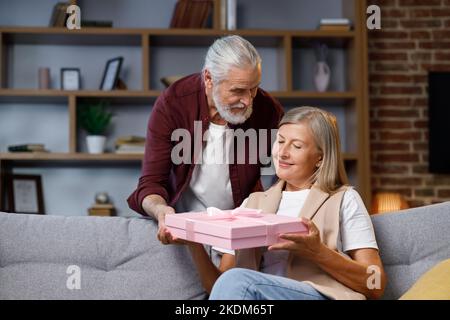 Premuroso vecchio marito dando romantico regalo di compleanno a moglie matura eccitato, amore uomo più anziano sorpresa felice donna di mezza età, scatola rosa aperta, bacio Foto Stock