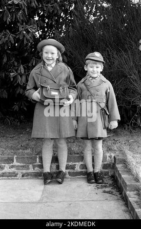 1963, storico, fratello e sorella stanno insieme per una foto fuori nella loro divisa scuola, cappotti e cappelli, la ragazza orgogliosamente tenendo la sua borsa a mano scuola, Inghilterra, Regno Unito, Foto Stock