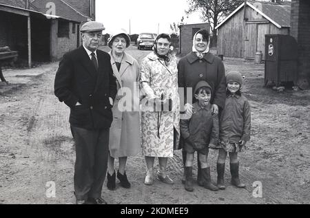 1960s, storico, in piedi insieme per una foto in un cortile, una famiglia, due nonni, una madre più la sua amica e due bambini piccoli, in una visita ad una fattoria, il ragazzo non impressionato..Tongue out,,,YUK! Inghilterra, Regno Unito. Foto Stock