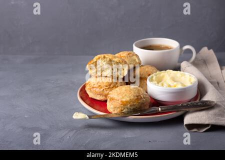 Focaccine appena sfornate fatte in casa con formaggio ed erbe su un piatto rosso con burro, una tazza di caffè su uno sfondo grigio testurizzato. Tradizionale bun inglese Foto Stock