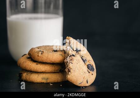 Biscotti classici con scaglie di cioccolato e bicchiere di latte su sfondo nero. Foto Stock