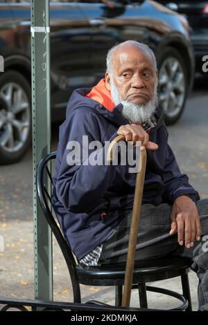 Un uomo più anziano, probabilmente musulmano, si siede e si appoggia sulla sua canna. Sulla 74th Street a Jackson Heights, Queens, New York City. Foto Stock