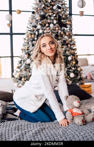 Una donna in una giacca bianca siede sul letto accanto all'albero di Natale. Natale Foto Stock