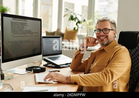 Un ingegnere IT maturo di successo che guarda la telecamera sul posto di lavoro mentre si siede davanti al monitor del computer e al laptop con dati codificati Foto Stock