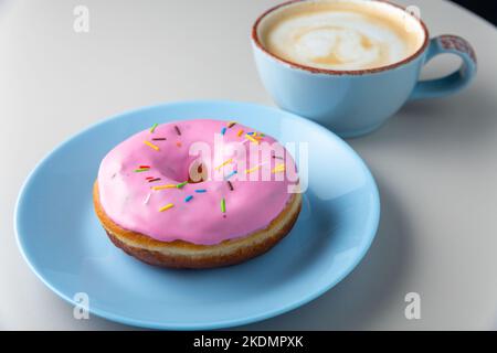 Ciambella ricoperta di smalto rosa e spolverate multicolore sul piatto blu e tazza di cappuccino con schiuma di latte su un tavolo bianco, vista normale. Spazio di copia Foto Stock