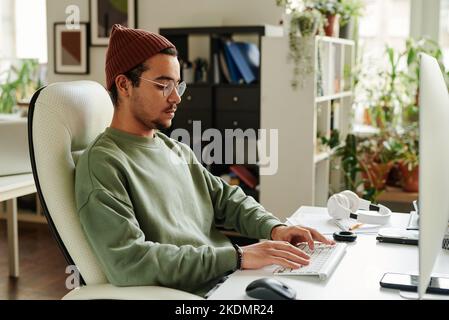 Serio ragazzo in casualwear a digitare sulla tastiera, mentre seduto di fronte al monitor del computer e di lavoro su un nuovo sito web o software Foto Stock