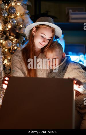 Adorabile ragazza caucasica felice e ragazzo di apertura di un regalo di Natale 2023. Allegro e allegro bambini che ricevono un regalo con decorato illuminato albero di Natale sullo sfondo. Immagine verticale di alta qualità Foto Stock