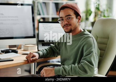 Giovane ingegnere INFORMATICO sicuro o sviluppatore di software in casualwear seduto in poltrona da monitor e computer portatile e guardando la fotocamera Foto Stock