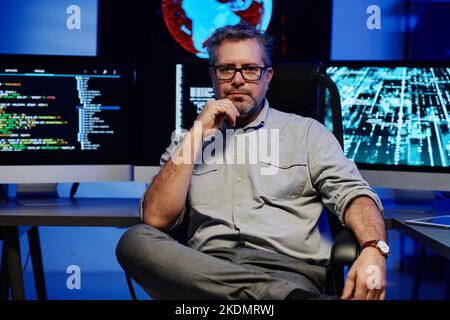 Serio sviluppatore di software maschile maturo in smart casualwear e occhiali seduti contro monitor di computer in sala server Foto Stock