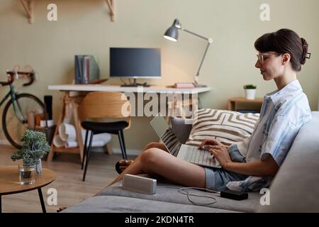 Ritratto con vista laterale di una giovane donna sorridente che utilizza un computer portatile mentre si rilassa sul divano in un ambiente accogliente, spazio copia Foto Stock