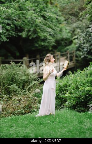 Donna che indossa un abito rosa che legge un libro in un giardino idilliaco Foto Stock