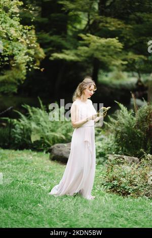 Donna che indossa un abito rosa che legge un libro in un giardino idilliaco Foto Stock