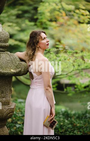 Donna che indossa un abito rosa che legge un libro in un giardino idilliaco Foto Stock