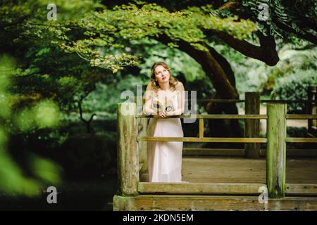 Donna che indossa un abito rosa che legge un libro in un giardino idilliaco Foto Stock