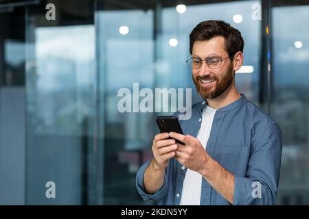 Uomo d'affari di successo in occhiali e barba in piedi vicino alla finestra di sera, uomo che utilizza il telefono, felice capo digitando il messaggio e navigando le pagine online sull'app per smartphone all'interno dell'ufficio. Foto Stock