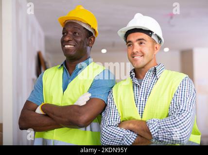 Ritratto di due ingegneri positivi in gilet giallo e casco di sicurezza in cottage ristrutturato Foto Stock