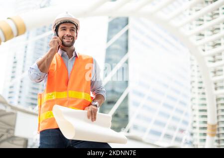 Gli ingegneri del Caucaso usano uno smartphone per parlare, indossando un giubbotto arancione e un grande elmetto, e l'altra mano che tiene la pianta bianca del pavimento. Foto Stock