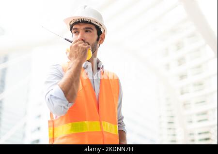 Gli ingegneri dell'uomo caucasico usano un walkie-talkie per parlare, indossando un giubbotto arancione e un grande elmetto nel lavoro sul sito del centro città. Foto Stock
