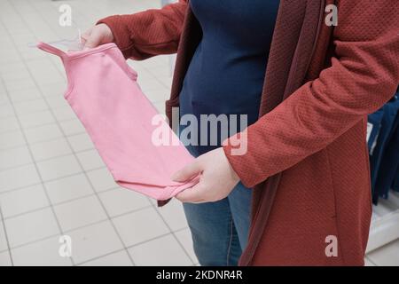 Donna incinta sceglie un vestito rosa - corpetto per un bambino in un negozio per acquistare Foto Stock