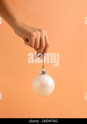 Composizione minima di Natale fatta di mano della donna che tiene bianco brillante Natale abbauble su sfondo arancione pastello. Felice Anno Nuovo. Foto Stock