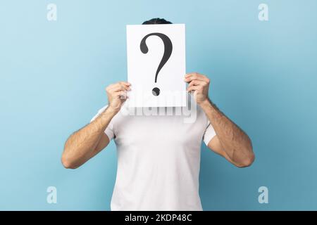 Ritratto dell'uomo che indossa una T-shirt bianca che nasconde il volto dietro un foglio bianco con punto interrogativo, trovare una soluzione intelligente, chiedere consigli. Studio in interni isolato su sfondo blu. Foto Stock