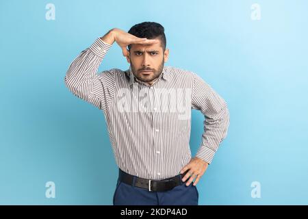 Ritratto di un bel uomo d'affari che guarda in lontananza, tiene la mano sugli occhi e guarda l'orizzonte con una vista attenta, indossando una camicia a righe. Studio in interni isolato su sfondo blu. Foto Stock