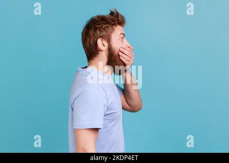 Non lo dico a nessuno. Vista laterale del bel giovane adulto bearded uomo zippando la bocca, mantenendo le informazioni segrete, guardando lo spazio laterale. Studio in interni isolato su sfondo blu. Foto Stock