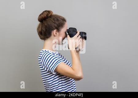 Vista laterale di una donna fotografa o di un viaggiatore che indossa una T-shirt a righe con fotocamera reflex digitale professionale e messa a fuoco, scatto di foto e ripresa di video. Studio in interni isolato su sfondo grigio. Foto Stock