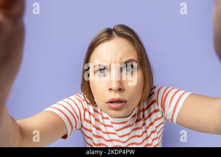 Ritratto di una donna bionda concentrata attenta che indossa una T-shirt a righe scattando foto al selfie, fissando la fotocamera dello smartphone, foto dal punto di vista. Studio al coperto isolato su sfondo viola. Foto Stock