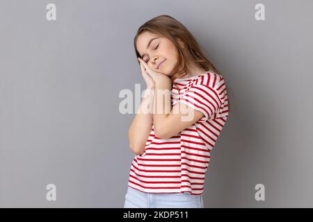 Ho bisogno di riposo. Ritratto di bambina che indossa una T-shirt a righe che sbadigna e copre la bocca con la mano, sentendosi esausta, mancanza di sonno. Studio in interni isolato su sfondo grigio. Foto Stock