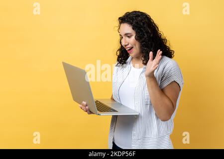 Ritratto di amichevole felice donna compiaciuta con capelli scuri ondeggianti lavorando sul portatile, videochiamare, agitare la mano, dire ciao o addio. Studio al coperto isolato su sfondo giallo. Foto Stock