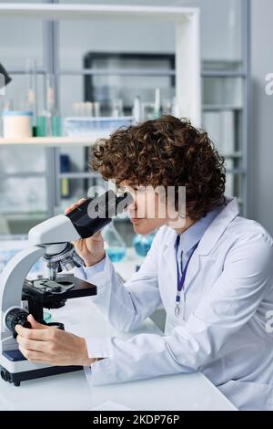 Giovane ricercatrice seria in camice guardando al microscopio mentre si siede sul posto di lavoro in laboratorio e studiando nuovo virus Foto Stock