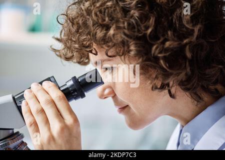 Vista laterale di una giovane biochimica femminile seria che guarda al microscopio durante la ricerca scientifica dal posto di lavoro in laboratorio clinico Foto Stock