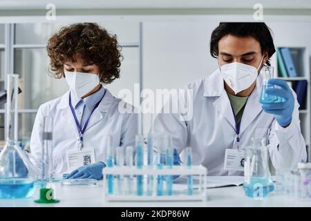 Due giovani medici in camici, respiratori e guanti che lavorano con sostanze chimiche liquide create in laboratorio durante la sperimentazione Foto Stock