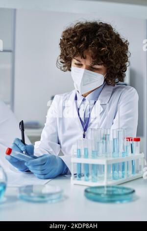 Giovane medico con camice da laboratorio, respiratore protettivo e guanti che scrivono il nome del campione mentre si siede sul posto di lavoro accanto a un set di matracci Foto Stock