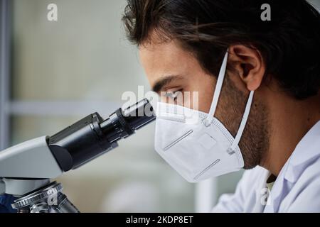 Primo piano di un giovane nel respiratore che guarda al microscopio in laboratorio durante un esperimento scientifico o uno studio di nuovi virus o batteri Foto Stock