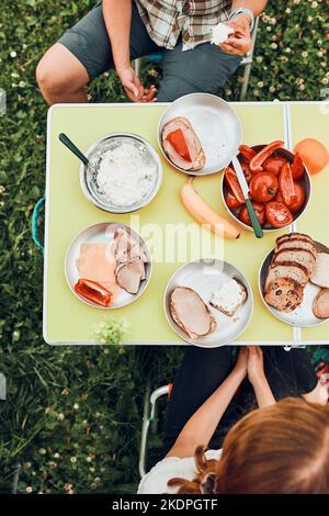 Famiglia che ha la colazione all'aperto in campeggio durante le vacanze estive. Pane, formaggio di ricotta, carne fredda, pomodori, frutta e tazze di caffè sul tavolo. Chiudi Foto Stock