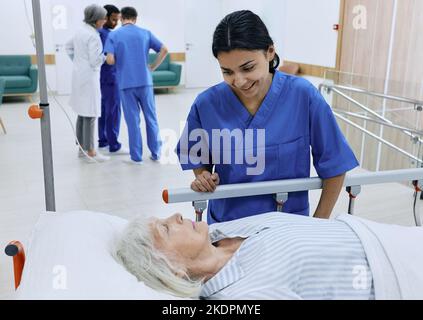 Infermiera positiva sostiene la donna anziana malata che giace sulla barella in corridoio ospedaliero sullo sfondo del consiglio dei medici. cura dei pazienti in ospedale Foto Stock