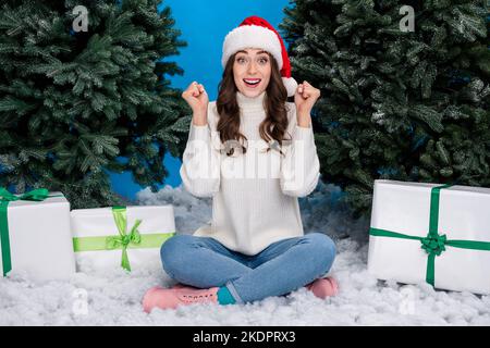 Foto della ragazza stupita fortunata vestita babbo natale helper cappello ottenere x-mas regali in aumento pugni isolato di colore blu sfondo Foto Stock