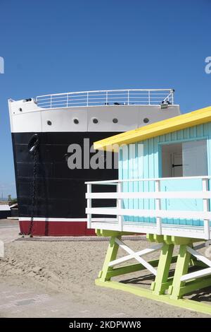 Port-Barcarès, Francia - 2022 ottobre; l'ex nave da crociera le Lydia sulla spiaggia accanto alla torre del bagnino dai colori vivaci sullo sfondo del cielo blu Foto Stock