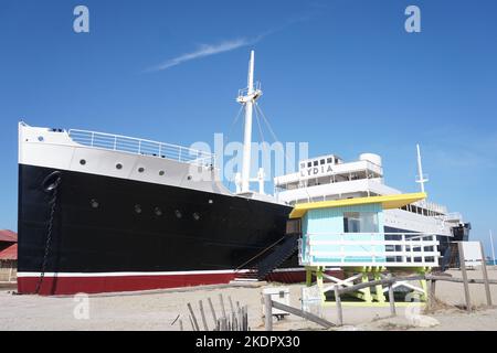 Port-Barcarès, Francia - 2022 ottobre; l'ex nave da crociera le Lydia sulla spiaggia accanto alla torre del bagnino dai colori vivaci sullo sfondo del cielo blu Foto Stock